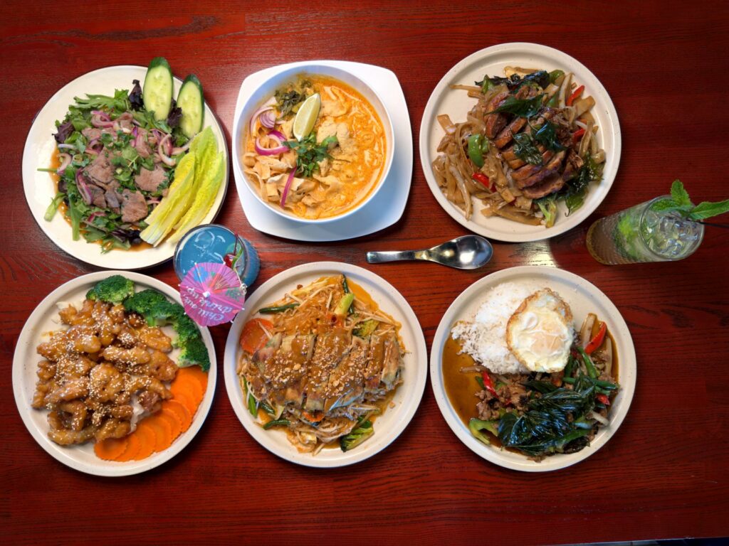 Six Thai dishes shared across one table at Nakhon Luang Thai Kitchen in Colorado Springs, including khao soi, roasted duck drunken noodles and pad gra pao