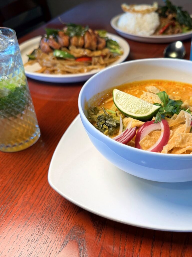 Khao soi in the foreground with roasted duck drunken noodles and pad gra pao behind it on a dining table