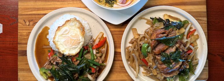 Pad gra pao with rice, roasted duck drunken noodles and a bowl of khao soi served together on a wooden board